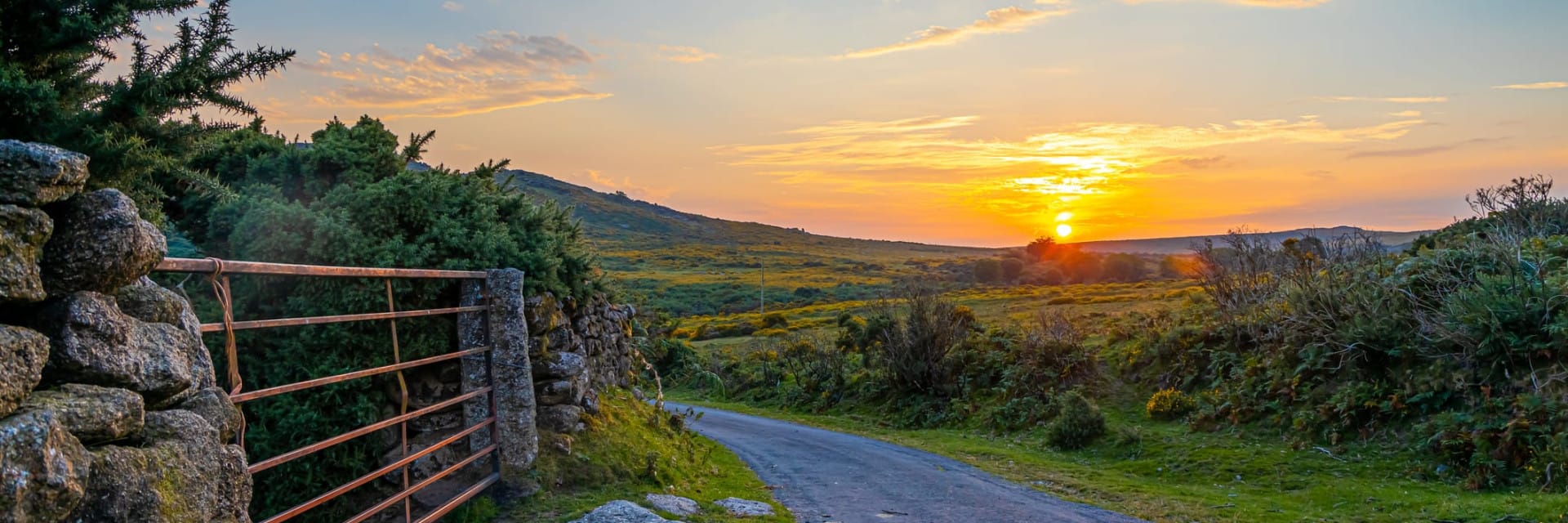 A scenic countryside road winds through green fields and shrubs at sunset, with a stone wall and metal gate on the left, and a colorful sky filled with clouds and sunlight.