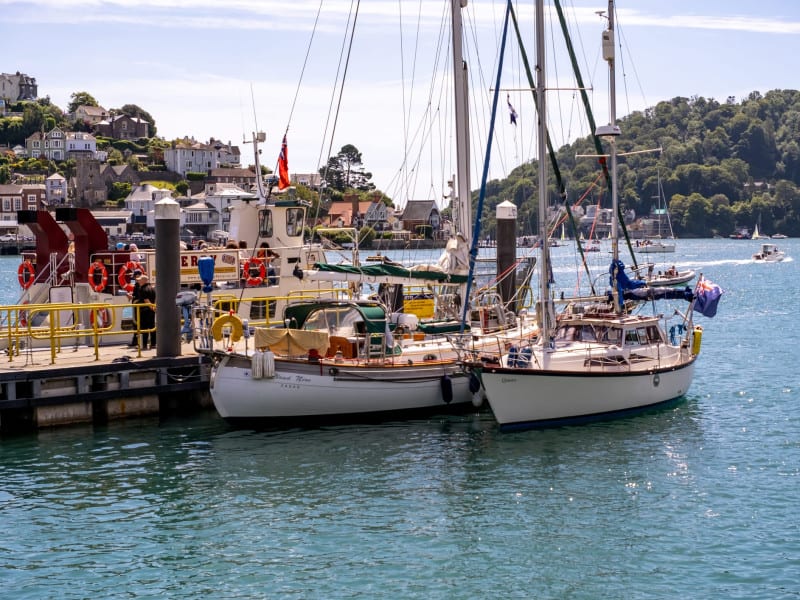 Sailboats and a small ferry are moored at a harbor with calm blue waters, surrounded by hills covered in trees and houses, under a sunny sky with boats visible in the distance.
