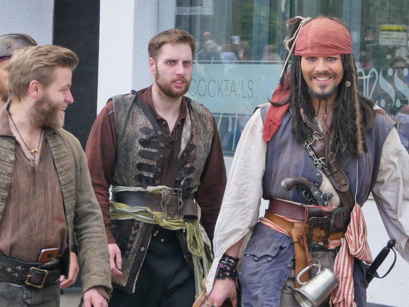 A group of people dressed as pirates walk together, wearing detailed costumes with hats, belts, and accessories, including one with a red bandana and long dreadlocks, smiling confidently.