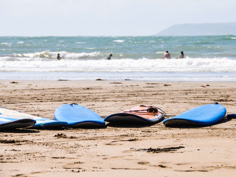 Surfboards lying on a sandy beach with gentle waves in the background, where people are swimming and enjoying the ocean on a sunny day.